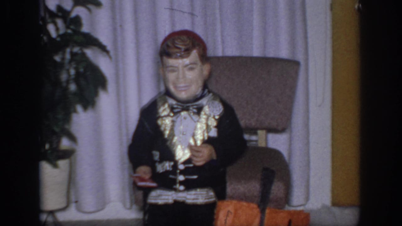 Ventriloquist dummy by curtains, chair, plant, and orange object.