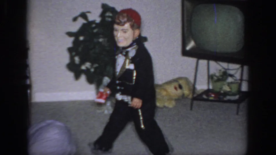 1966 Sacramento home-movie frame showing the dummy in profile on a carpeted living room floor near a houseplant, tube television, and toy-like object.
