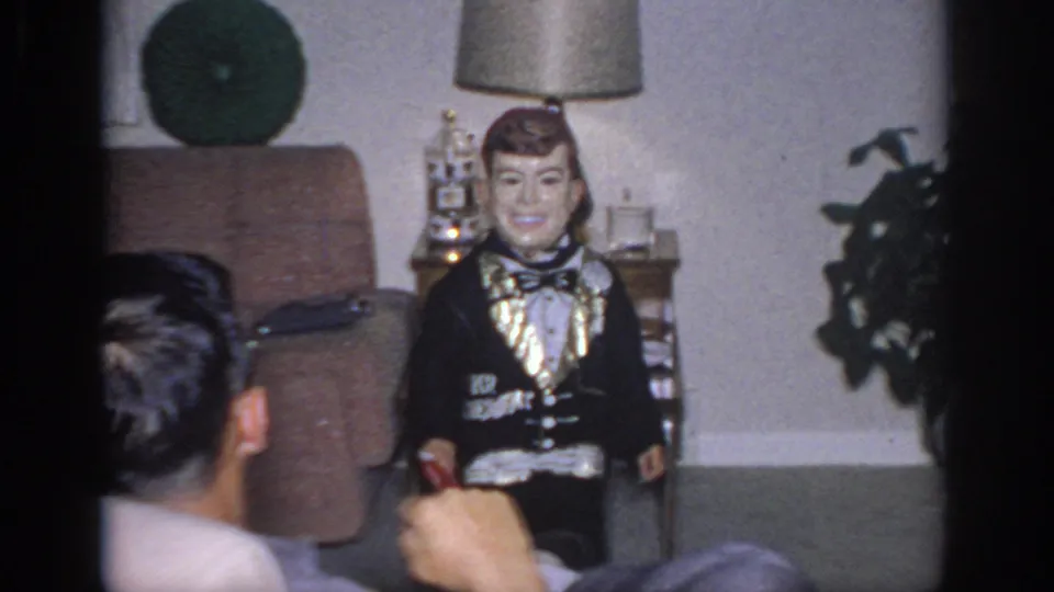 1966 Sacramento frame showing the dummy near a chair and lamp while an adult head and hand are partly visible in the foreground.