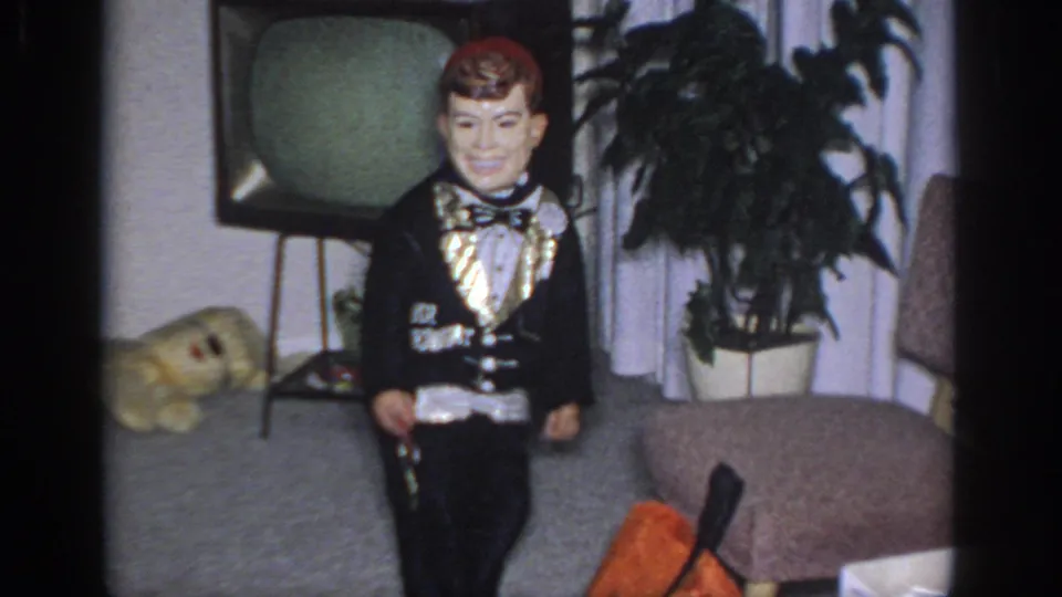 1966 Sacramento home-movie frame showing a costumed man in a living room with a television, curtains, potted plant, and sofa.