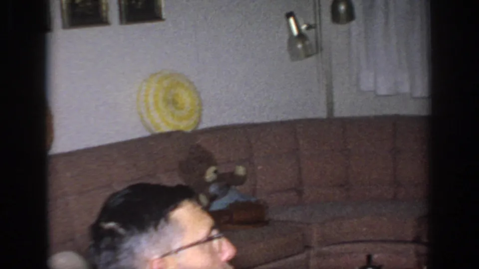 1966 Sacramento home-movie frame showing a sofa, yellow circular pillow, doorway, wall art, and a partially visible man with glasses.