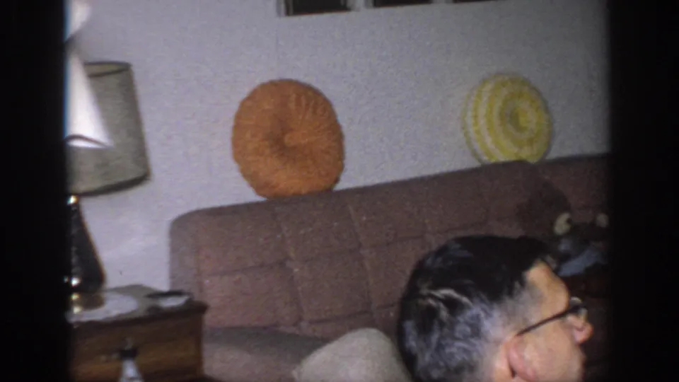 1966 Sacramento interior frame with a brown sofa, orange and yellow pillows, table lamp, side table, and partial profile of a man.