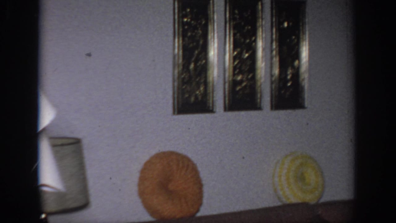 1966 Sacramento living room frame showing framed wall art, brown sofa, orange and yellow round pillows, and part of a lamp shade.