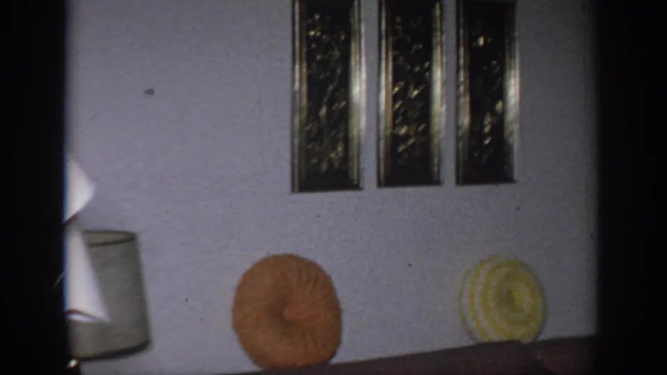 1966 Sacramento living room frame showing framed wall art, brown sofa, orange and yellow round pillows, and part of a lamp shade.