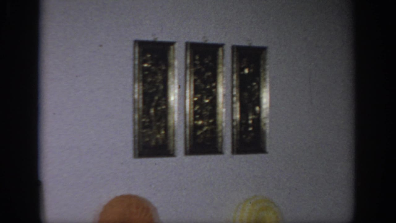 1966 Sacramento home-movie frame showing a textured living-room wall, three vertical gold-framed artworks, and partial circular pillows.