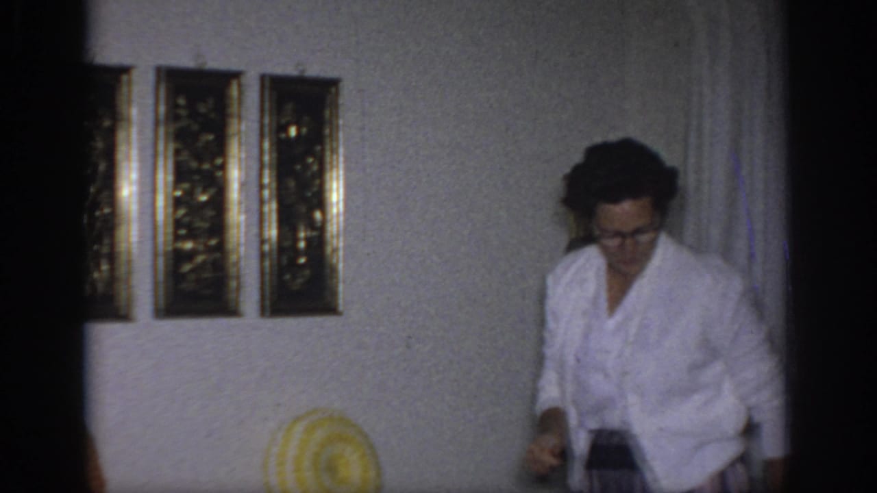 1966 Sacramento home-movie frame with a woman in glasses and a white cardigan entering a living room near framed wall art.