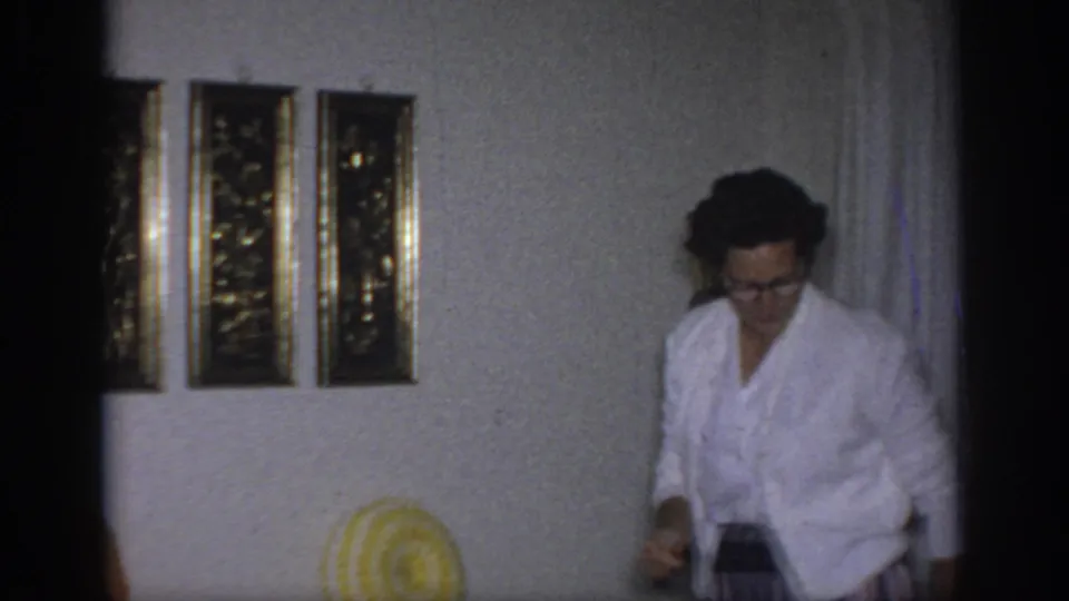 1966 Sacramento home-movie frame with a woman in glasses and a white cardigan entering a living room near framed wall art.