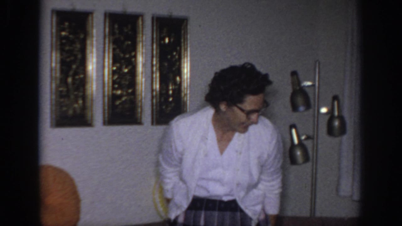 1966 Sacramento home-movie frame showing a woman in glasses moving through a living room near a multi-head floor lamp.