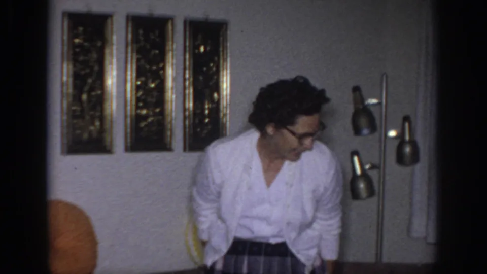 1966 Sacramento home-movie frame showing a woman in glasses moving through a living room near a multi-head floor lamp.