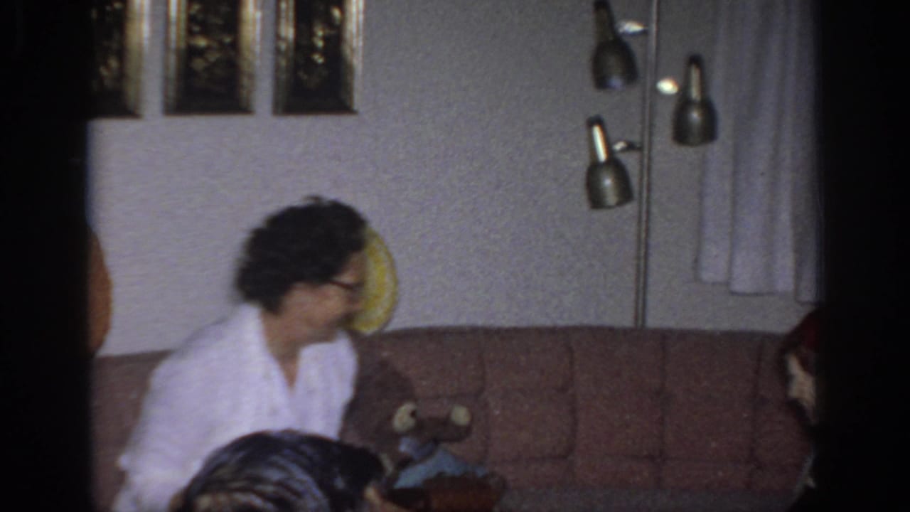 1966 Sacramento home-movie frame showing a woman sitting on a brown couch in a living room as another person appears partly at right.