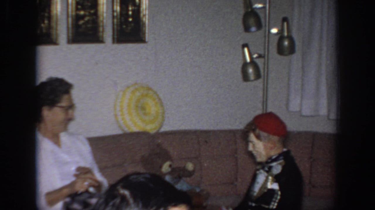 Woman in a white blouse sits on a couch beside a costumed ventriloquist dummy.