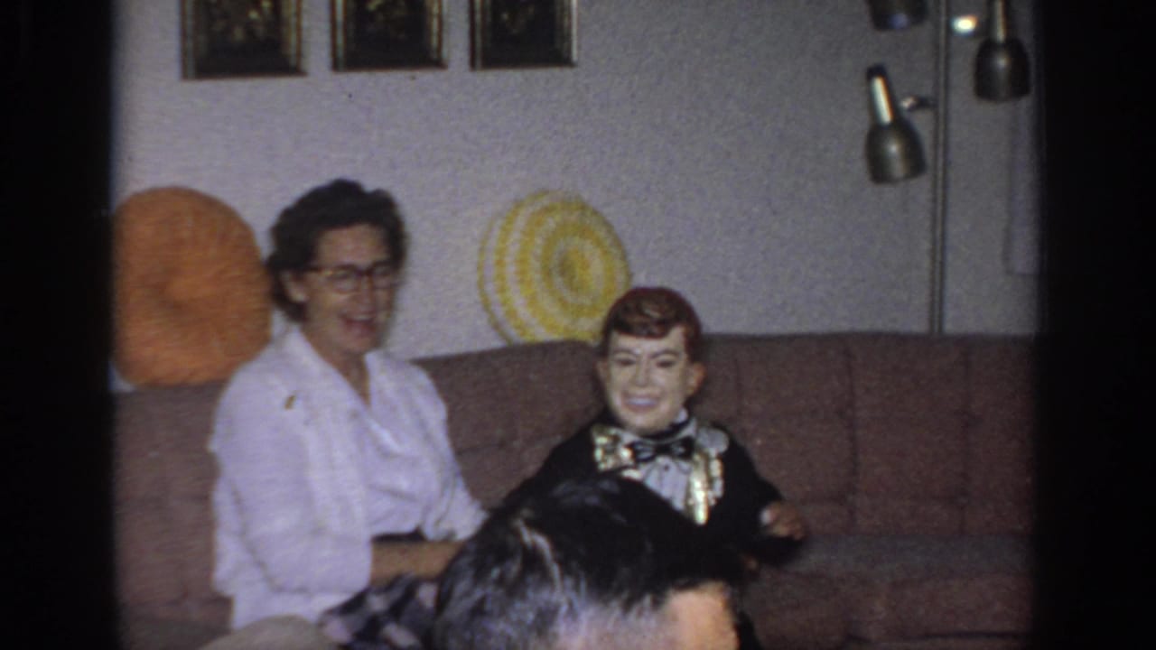 1966 Sacramento home-movie frame with a smiling woman and a tuxedoed ventriloquist dummy seated on a brown couch.