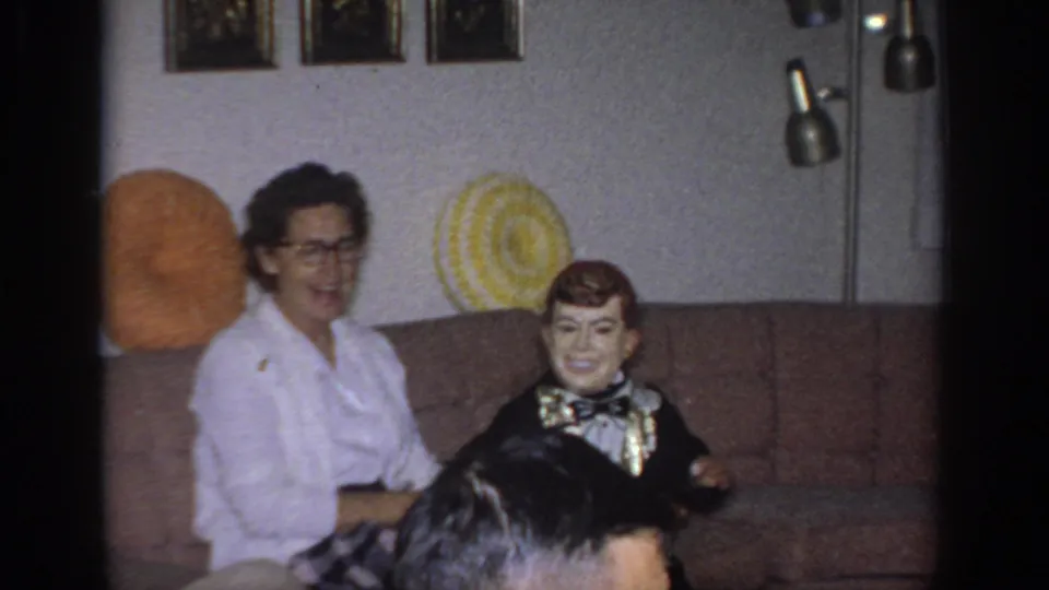 1966 Sacramento home-movie frame with a smiling woman and a tuxedoed ventriloquist dummy seated on a brown couch.