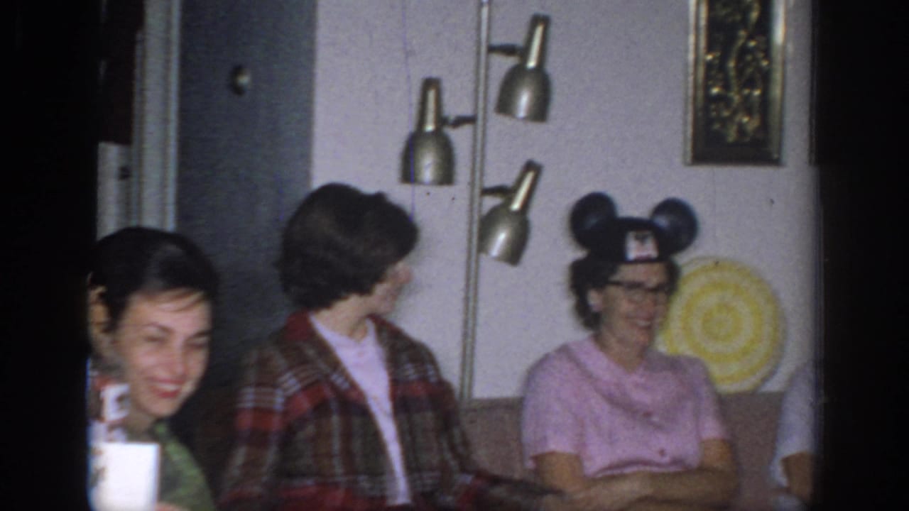1966 Sacramento home-movie frame showing women seated indoors, including one in Mickey Mouse ears and another in plaid outerwear.