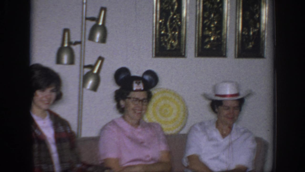 1966 Sacramento home-movie frame with women seated in a private living room, wearing novelty Mickey Mouse ears and a white hat.