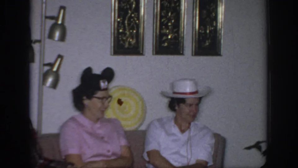 1966 Sacramento home-movie frame showing two women seated in a living room with novelty hats and mid-century wall decor.