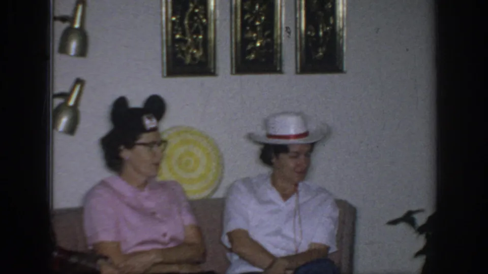 1966 Sacramento home-movie frame of two women sitting indoors, with a Mickey Mouse ears hat and a white cowboy-style hat visible.