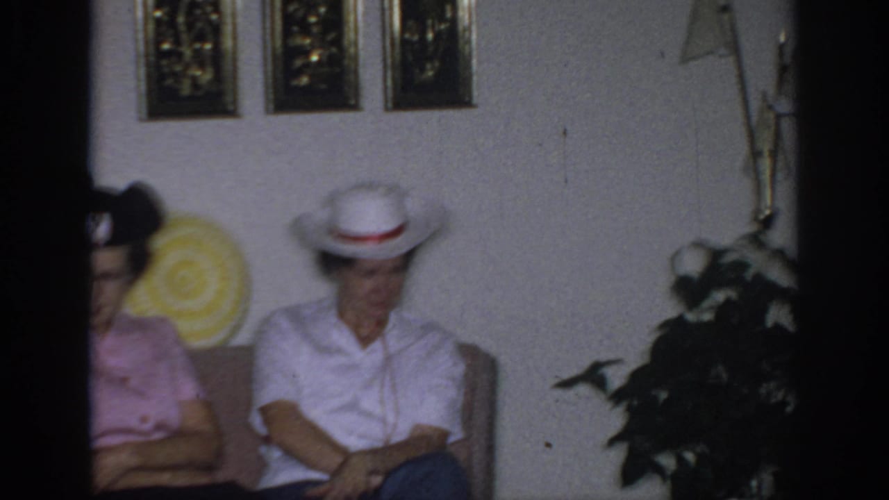 1966 Sacramento home-movie frame showing two women seated on a couch in a living room with wall art and a potted plant nearby.