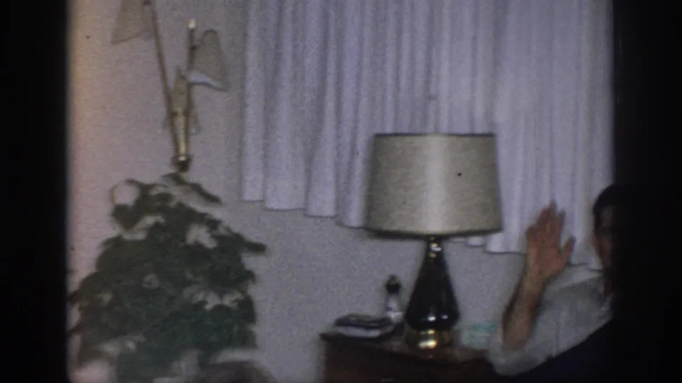 1966 Sacramento home-movie frame showing a seated adult raising a hand beside curtains, a table lamp, side table, and potted plant.