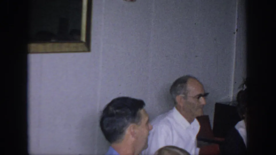 1966 Sacramento home-movie frame showing adults seated in a living room, including an older man with glasses and framed wall art behind them.