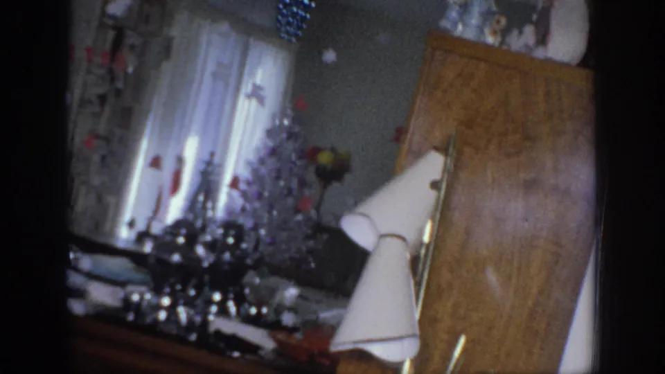 1966 Sacramento home-movie frame showing a decorated living room corner with curtains, tabletop holiday items, lamp, and cabinet.