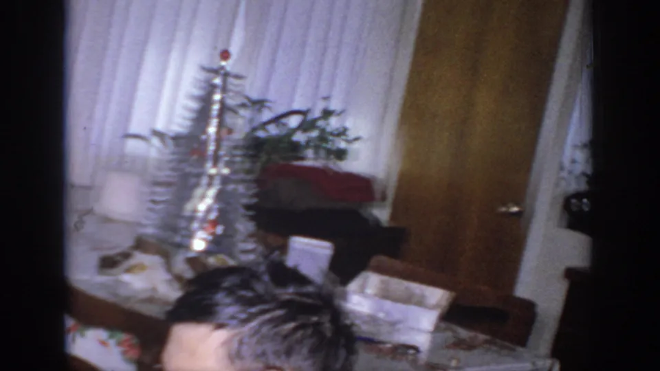 1966 Sacramento home-movie frame with silver tinsel tree, white curtains, wrapped boxes, furniture, and a partial person visible.