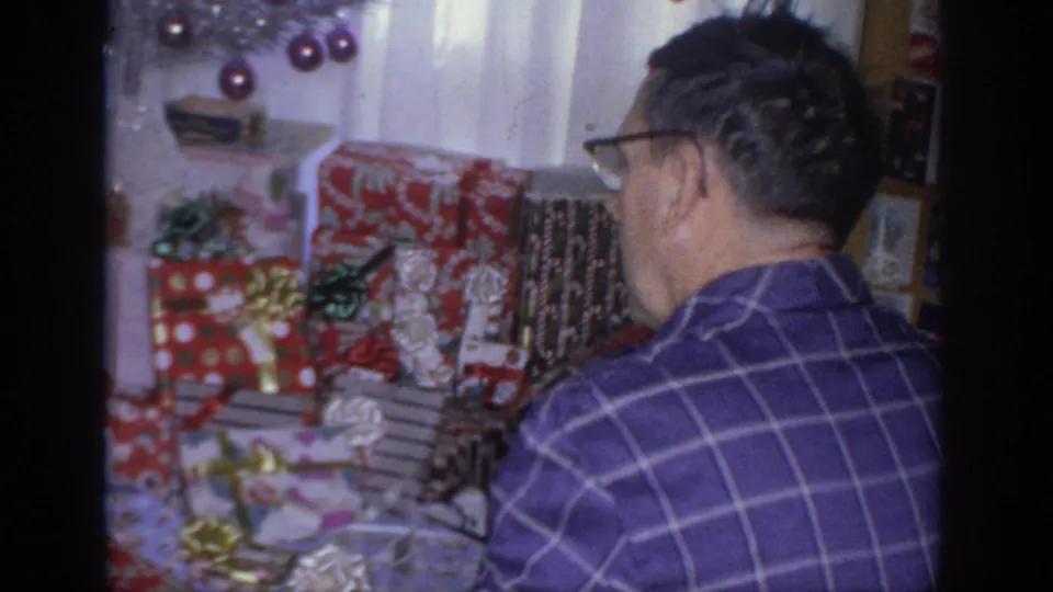1966 Sacramento frame with an older man in glasses near red and patterned wrapped Christmas gifts inside a decorated home.