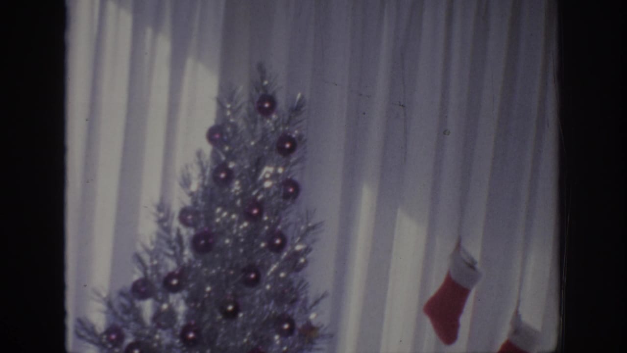 Silver Christmas tree near white curtains with two red stockings at the right.