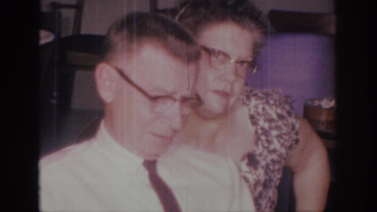 1966 Sacramento home-movie frame showing a man in a white shirt and dark tie beside a woman in a patterned dress indoors.