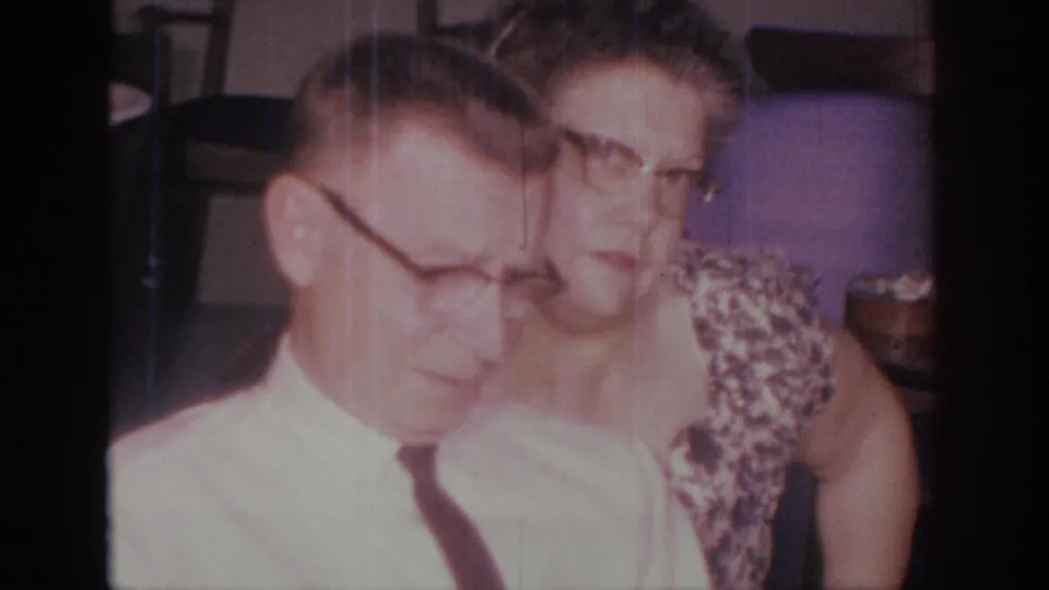 1966 Sacramento home-movie frame showing a man in a white shirt and dark tie beside a woman in a patterned dress indoors.