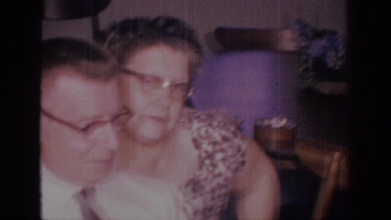 Close view of a seated man and woman with glasses in a dim domestic interior.