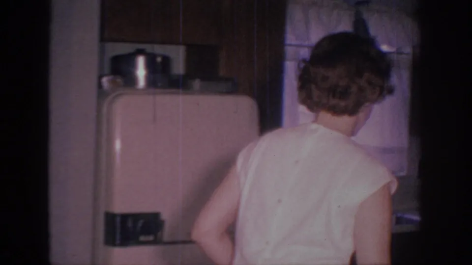 1966 Sacramento home-movie frame showing a woman from behind near a refrigerator and cabinets in a private kitchen.