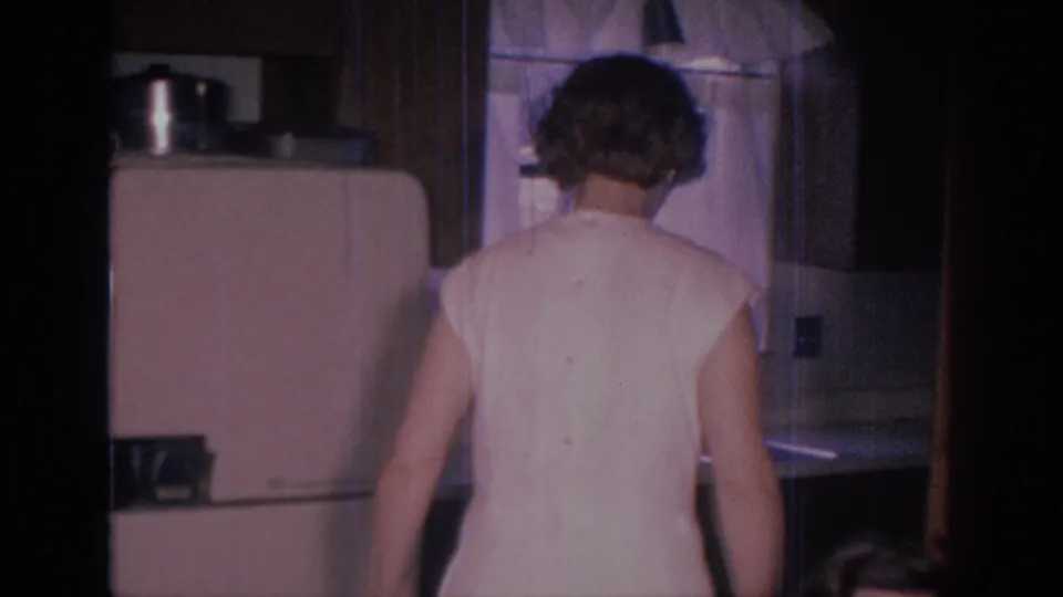 1966 Sacramento home-movie frame showing a woman from behind in a mid-century kitchen with refrigerator, counter, and sink area nearby.