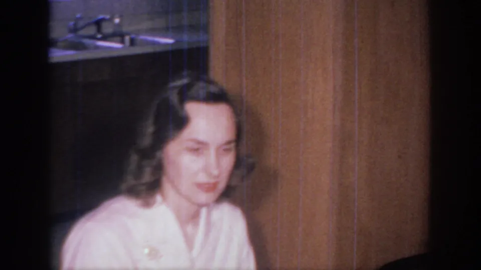 1966 Sacramento home-movie frame showing a woman in a light garment near wood paneling, with part of the kitchen sink and counter behind her.