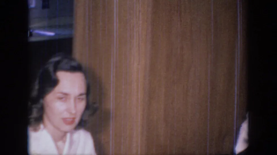 1966 Sacramento home-movie frame with a woman near wood paneling, looking off-camera as a partial figure appears at the right edge.