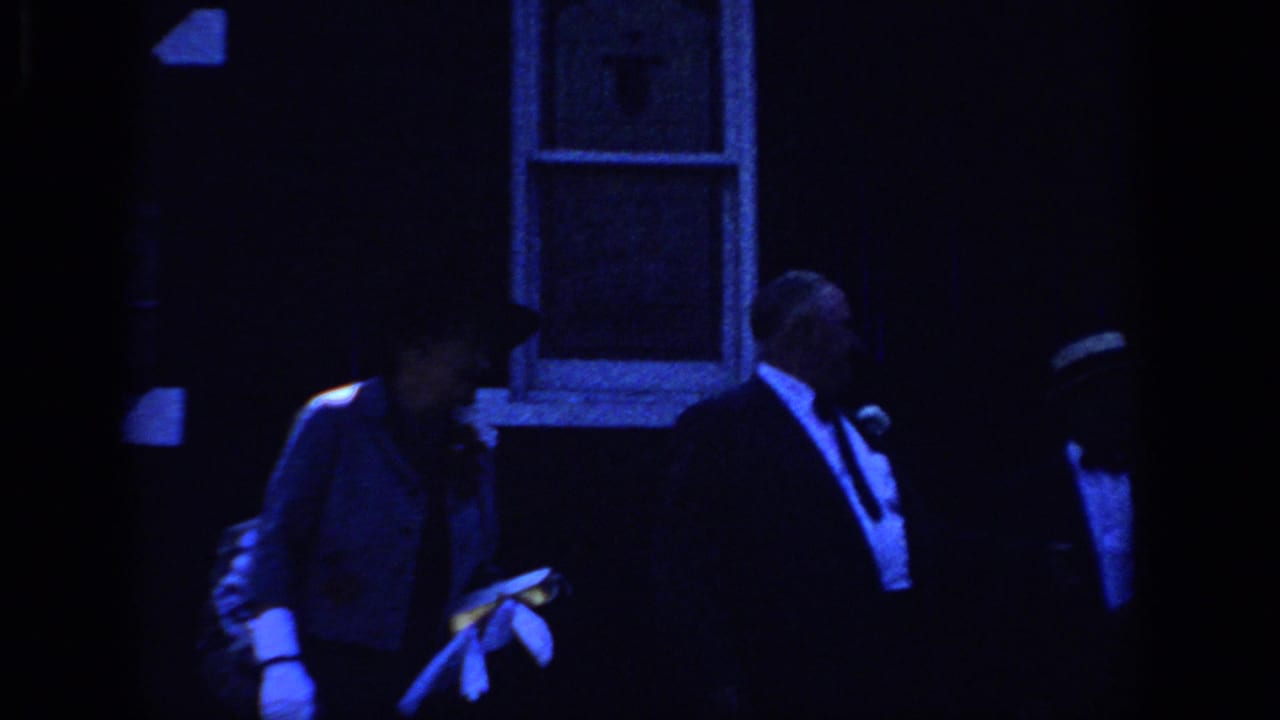 1966 Sacramento frame with a man in a tuxedo and a gloved woman near building windows during a dim evening gathering.