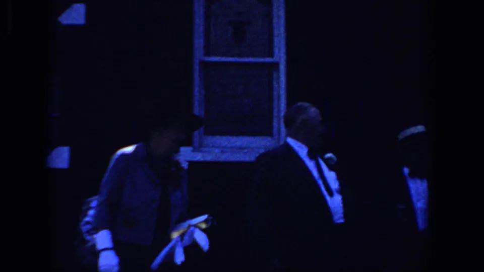 1966 Sacramento frame with a man in a tuxedo and a gloved woman near building windows during a dim evening gathering.