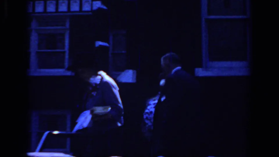 Blue-shifted 1966 Sacramento home-movie frame of formally dressed adults moving past a shadowed building exterior.
