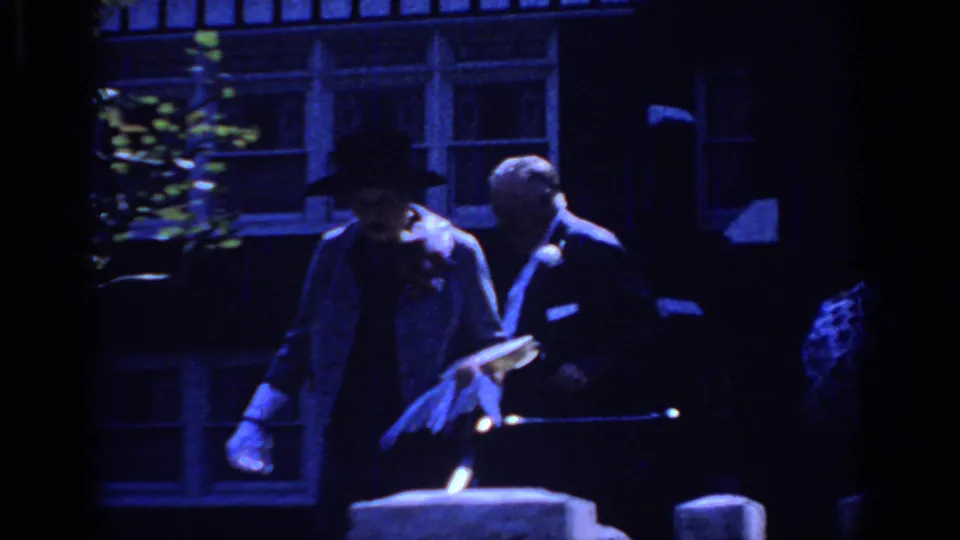 1966 Sacramento party frame showing a tuxedoed man beside a gloved guest near windows, railing, and dark exterior architecture.