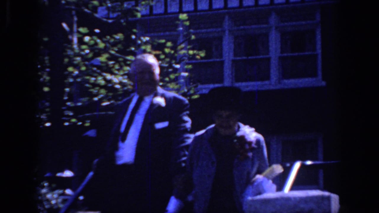 A man in a dark suit and a woman in a hat walk together past a house exterior.