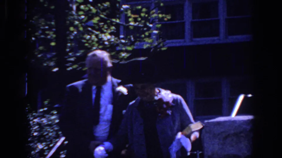A blue-cast 8mm frame shows the couple passing in front of windows and foliage during a formal social departure.