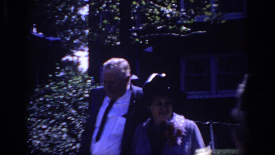 The man and woman continue walking outside the Sacramento home, with strong shadows, greenery, and house details visible behind them.