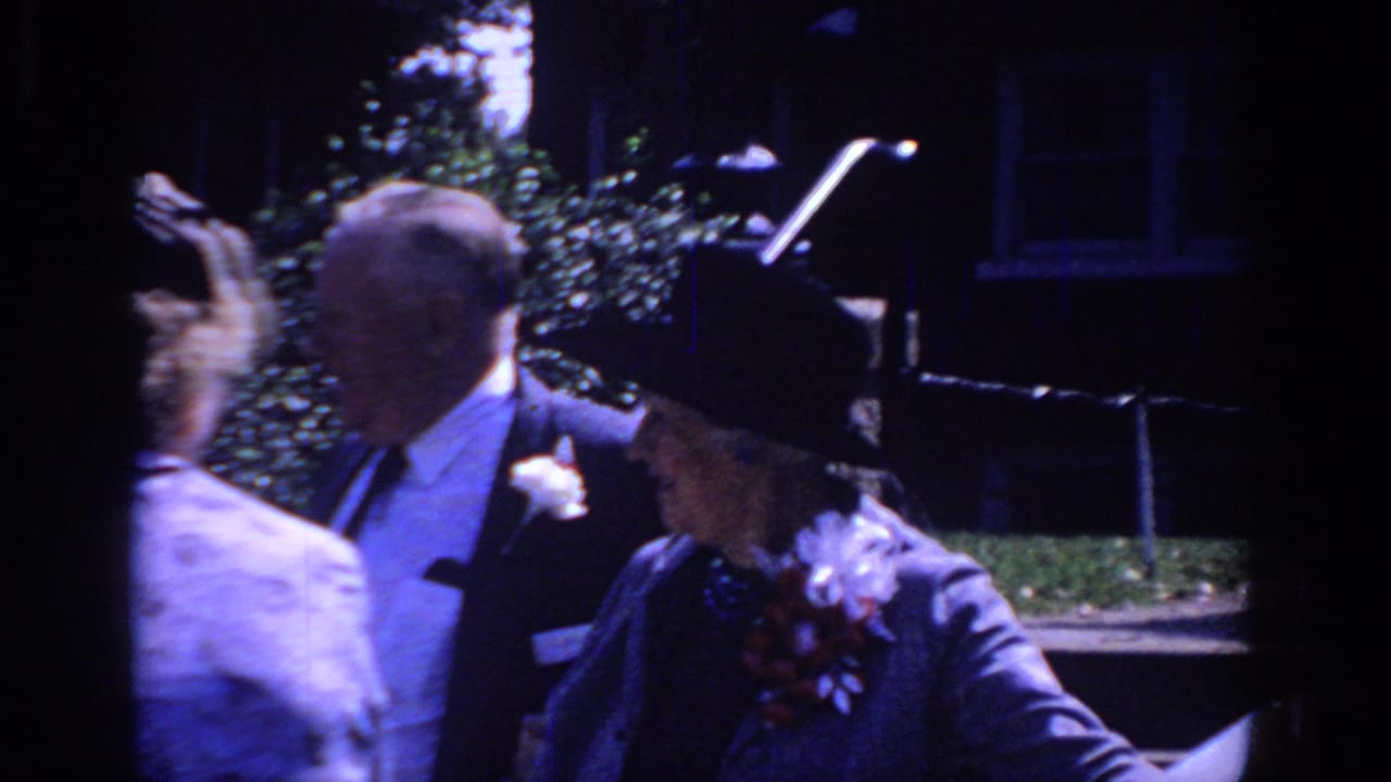 Sacramento 1966 home-movie frame showing formally dressed older adults near a stone wall, with hats, suits, and floral accessories visible.