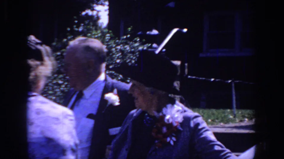 Sacramento 1966 home-movie frame showing formally dressed older adults near a stone wall, with hats, suits, and floral accessories visible.