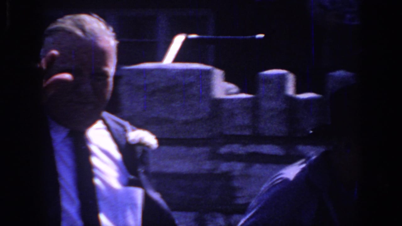 Sacramento 1966 frame of an older man in a suit and boutonniere, partly shadowed, standing near a stone wall during a formal gathering.