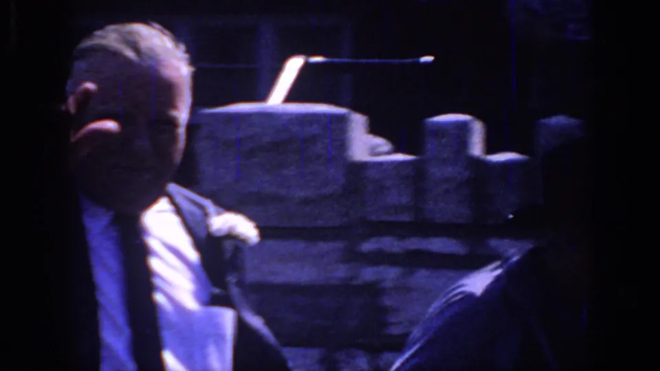 Sacramento 1966 frame of an older man in a suit and boutonniere, partly shadowed, standing near a stone wall during a formal gathering.