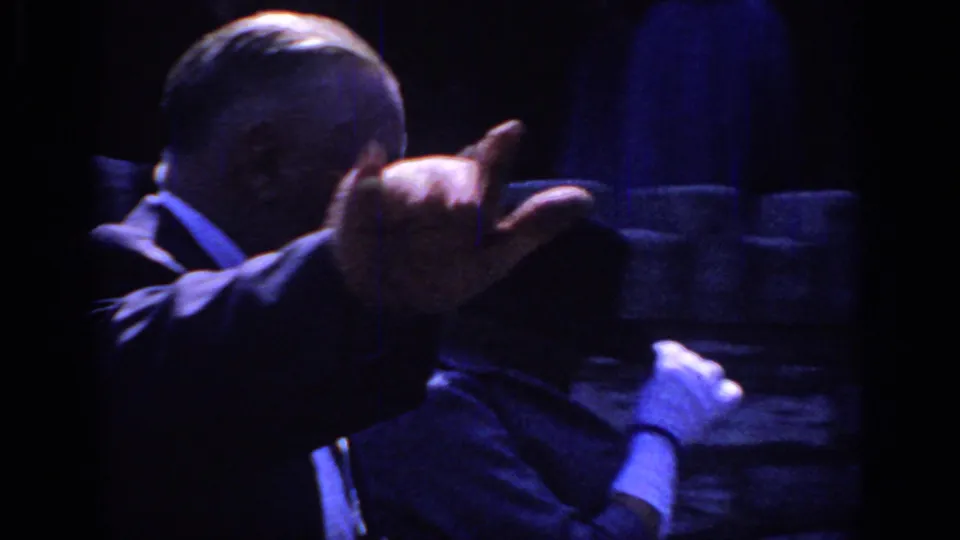 Sacramento 1966 home-movie frame showing an older man gesturing near a woman in a dark hat and gloves beside a stone wall.