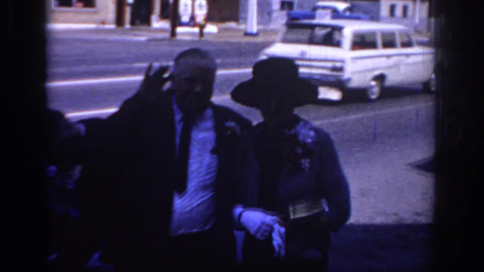 Sacramento 1966 frame showing an older couple in formal clothing near a street, with a white station wagon visible in the background.