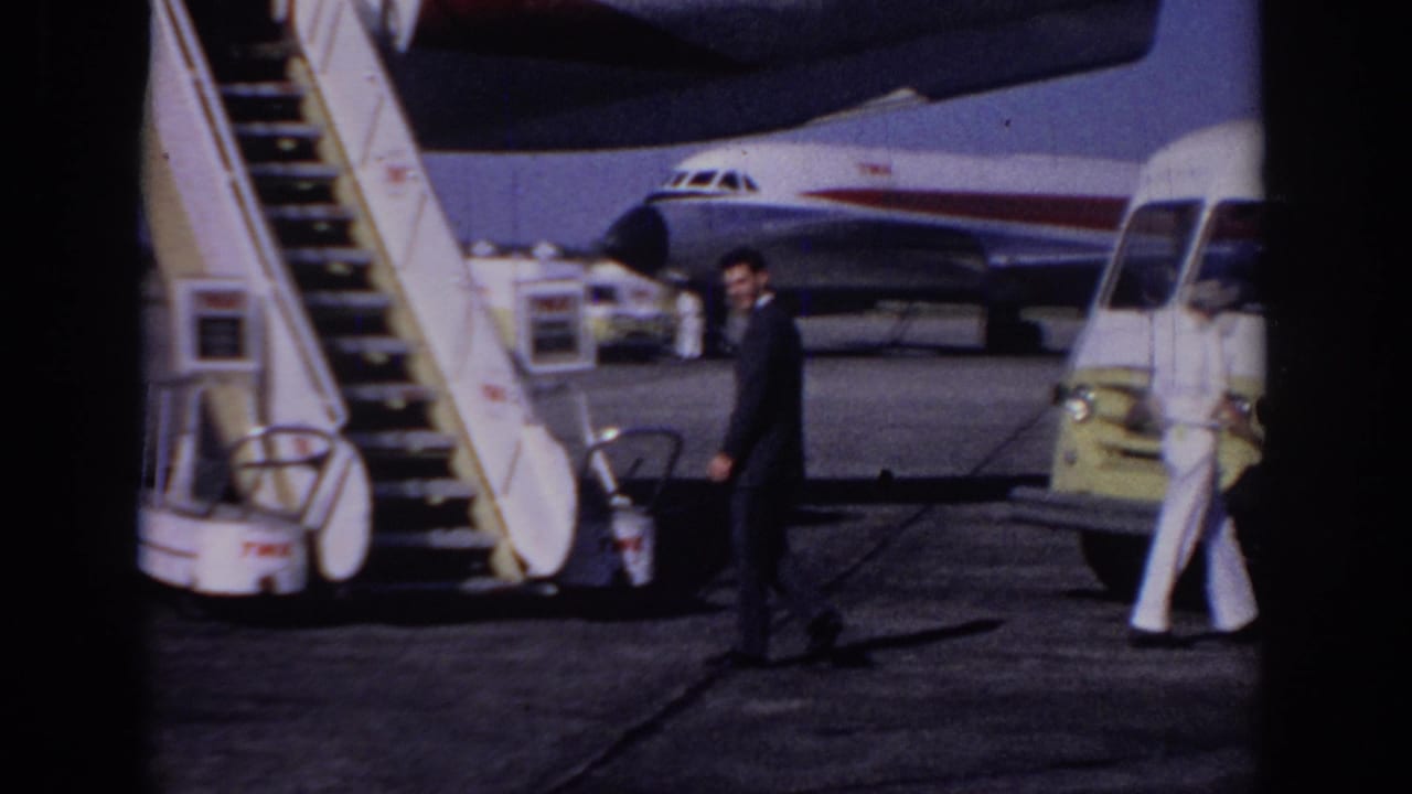 1966 Sacramento airport tarmac view with a suited man walking past mobile stairs, a yellow service van, and a TWA aircraft.