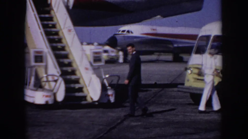 1966 Sacramento airport tarmac view with a suited man walking past mobile stairs, a yellow service van, and a TWA aircraft.